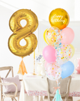 A balloon arrangement featuring a gold-colored number '8' balloon, along with rainbow balloons, tied together with ribbons. Balloons are in front of a dining table and chairs
