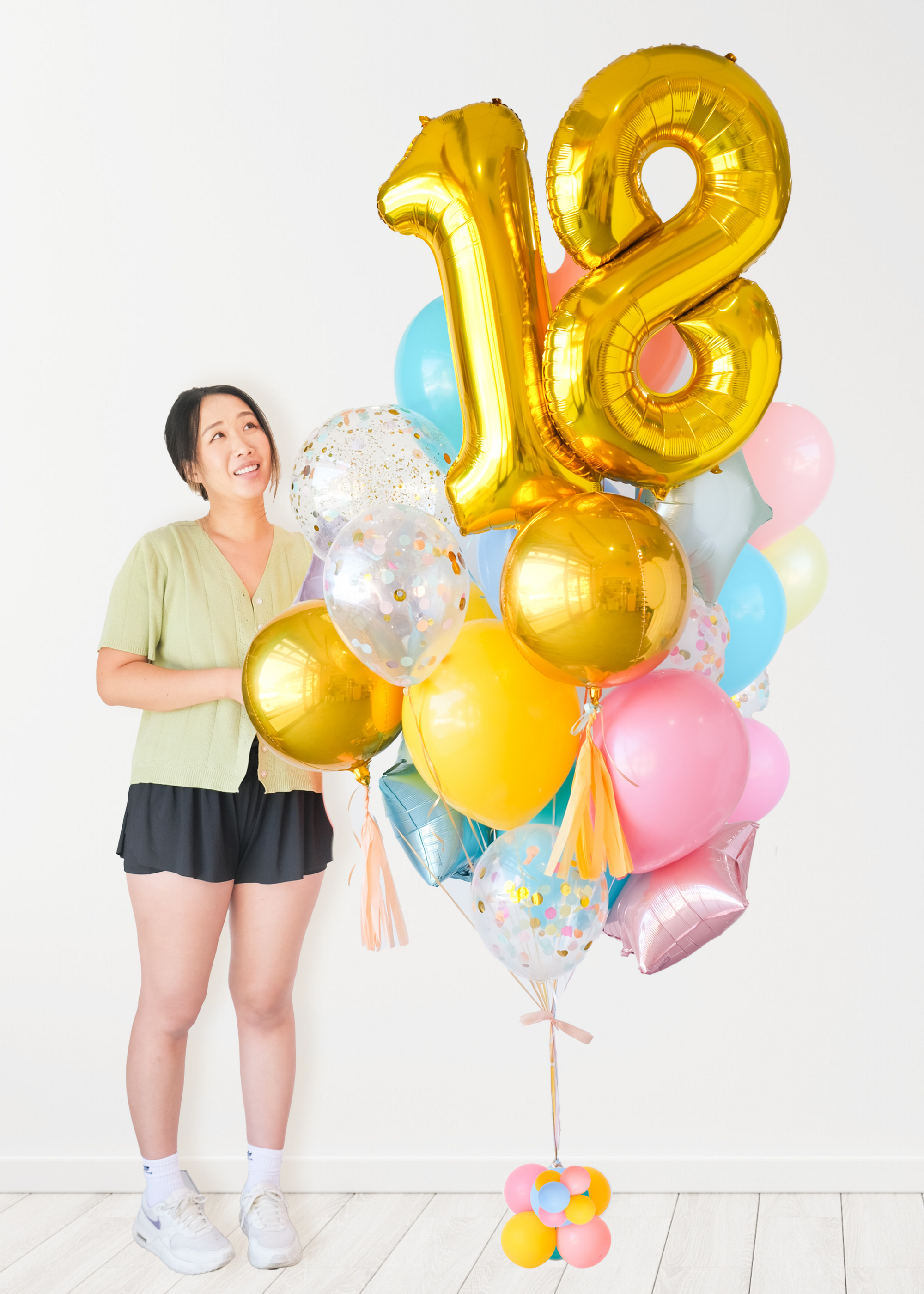 A balloon arrangement featuring a golden number '18' balloon with multicolored balloons tied to it, with the word 'Happy Birthday' printed on a gold balloon. A girl is standing next to it and look towards the bouquet.