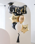 A balloon bouquet with black, gold, and silver balloons, including one that reads 'Grad' and star-shaped ones in front of cream backdrops