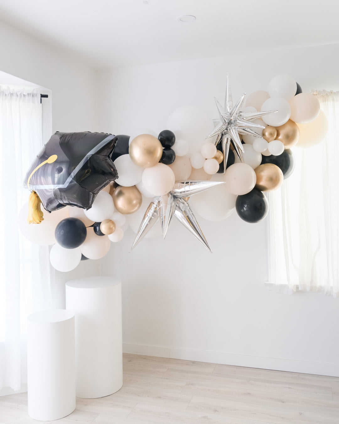 A balloon garland featuring a graduation cap design, with a mix of gold, silver, and white balloons, displayed indoors.