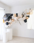 A balloon garland featuring a graduation cap design, with a mix of gold, silver, and white balloons, displayed indoors.