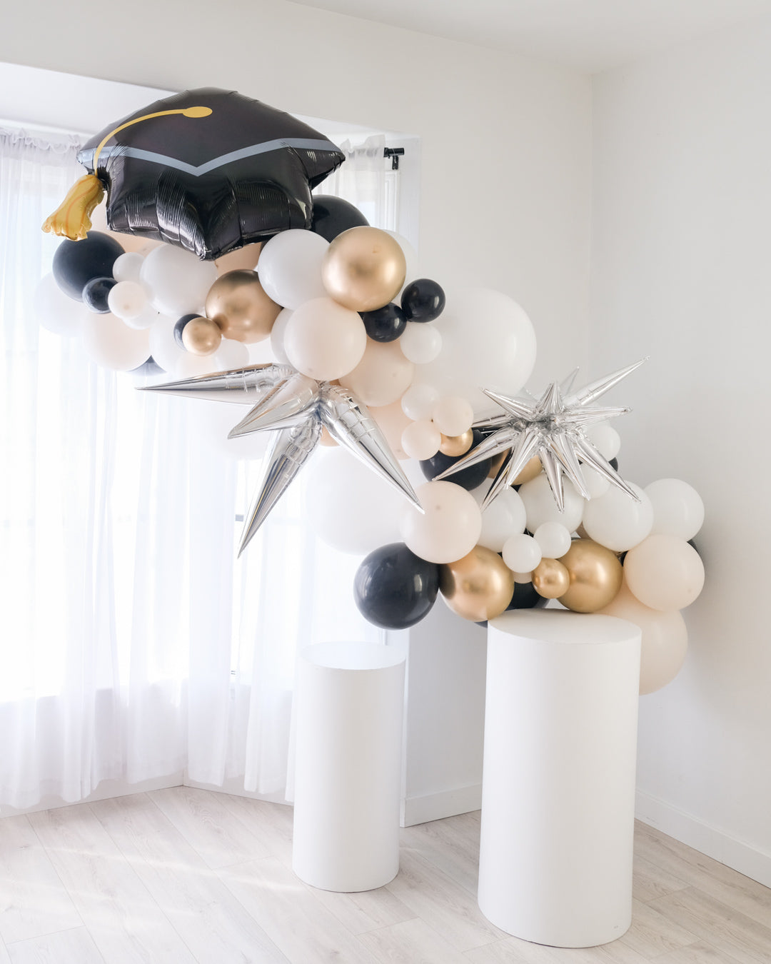 A balloon garland featuring a graduation cap design, with a mix of gold, silver, and white balloons, displayed indoors.