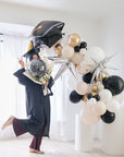 A balloon garland featuring a graduation cap design, with a mix of gold, silver, and white balloons, displayed indoors.