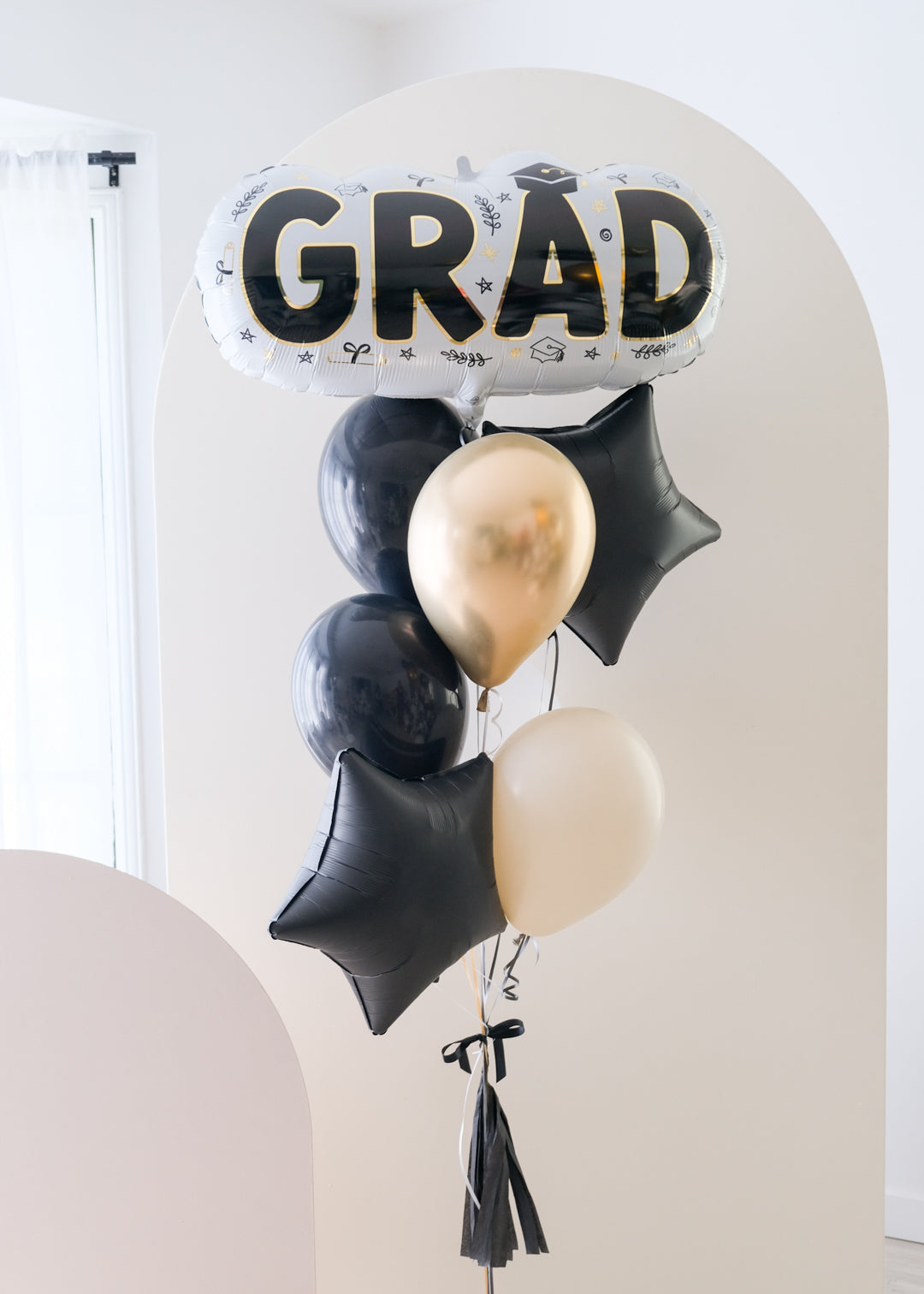 A balloon arrangement with a large 'GRAD' letter balloon in gold and black, surrounded by smaller black balloons, tied together with a signature bow, tassel, and bobble balloon weight.