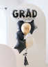 A balloon arrangement with a large 'GRAD' letter balloon in gold and black, surrounded by smaller black balloons, tied together with a signature bow, tassel, and bobble balloon weight.