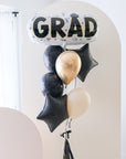 A balloon arrangement with a large 'GRAD' letter balloon in gold and black, surrounded by smaller black balloons, tied together with a signature bow, tassel, and bobble balloon weight.