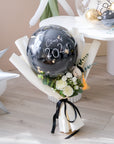 A mini graduation bouquet with a large black balloon featuring 'Class 2024, wrapped with black and white paper, and decorated with white and yellow flowers and a small black bear figurine.