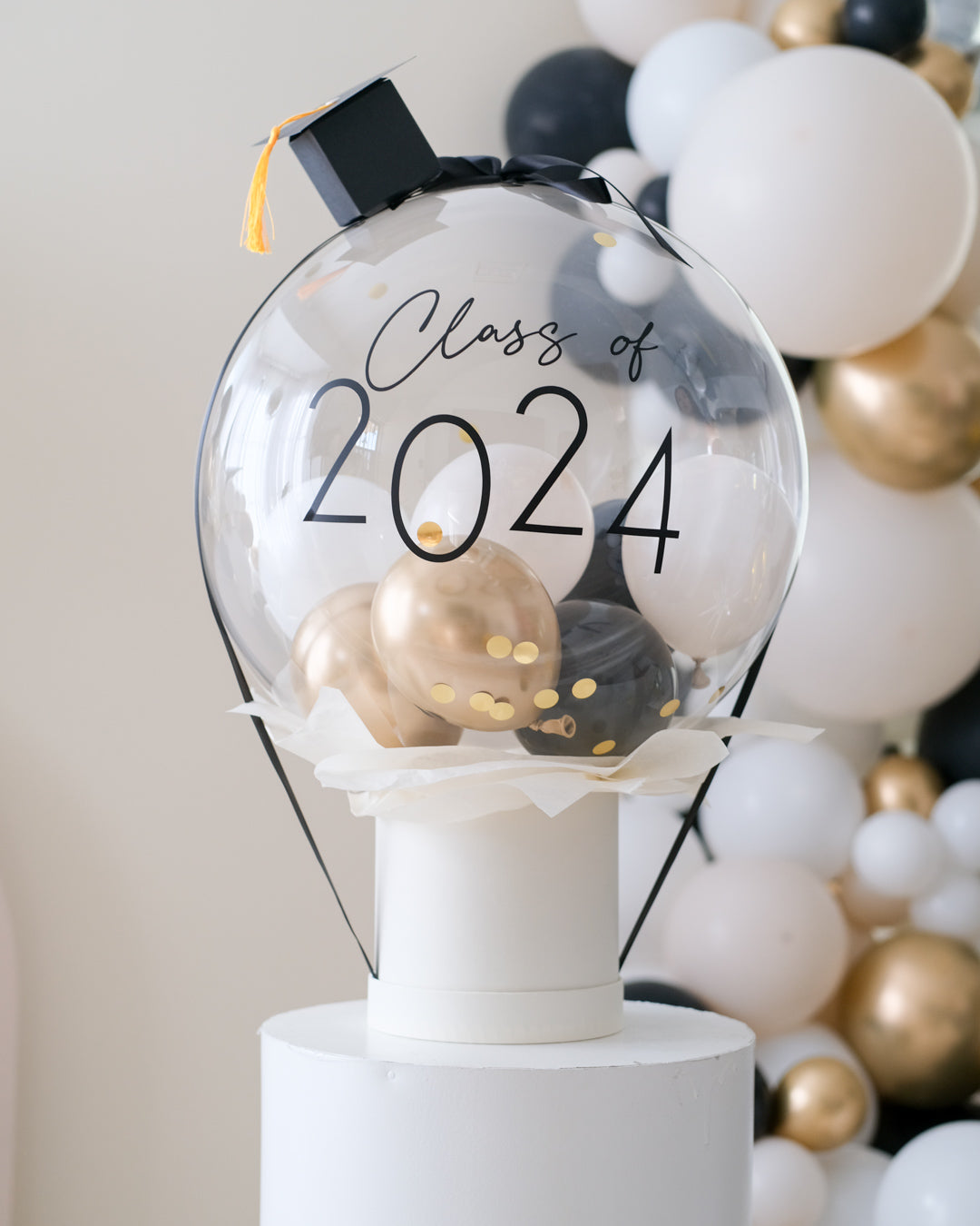 A clear balloon box with 'Class of 2024' written on it, containing gold and black mini balloons, displayed on a stand with a blurred background of similar balloons.