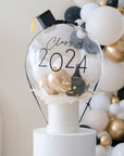 A clear balloon box with 'Class of 2024' written on it, containing gold and black mini balloons, displayed on a stand with a blurred background of similar balloons.
