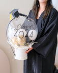 A decorative bubble balloon box with 'Class of 2025' printed on it, filled with multicolored balloons, mounted on a black stand.