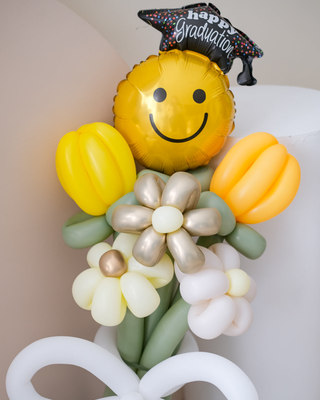 A balloon bouquet featuring a smiling face balloon, yellow and white flower-shaped balloons, and a balloon bow,