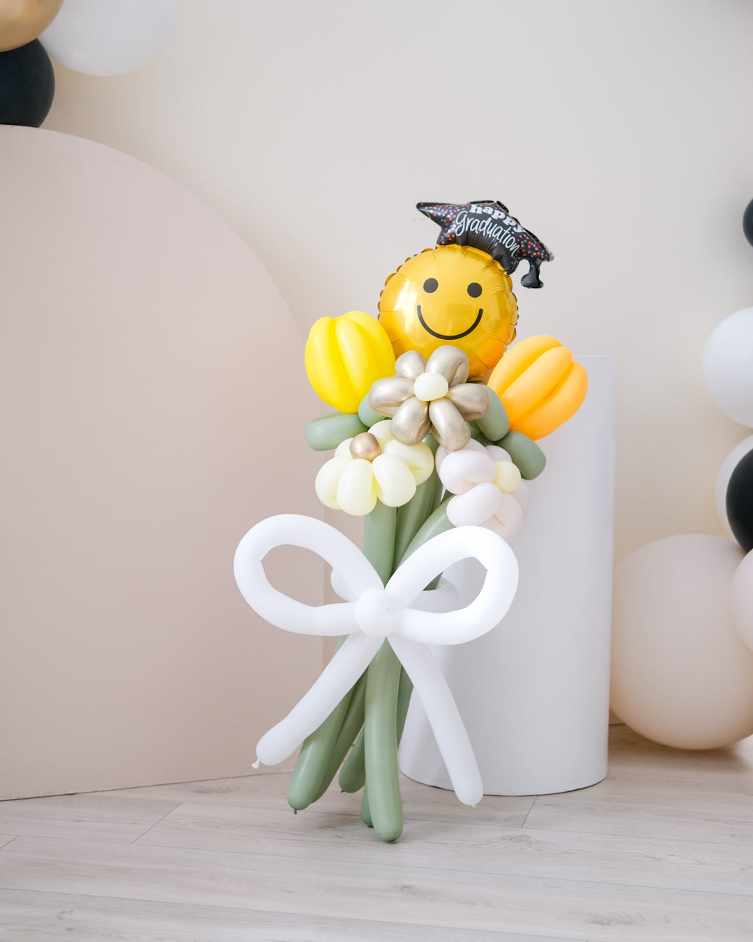 A balloon bouquet featuring a smiling face balloon, yellow and white flower-shaped balloons, and a balloon bow, in front of cream backdrops