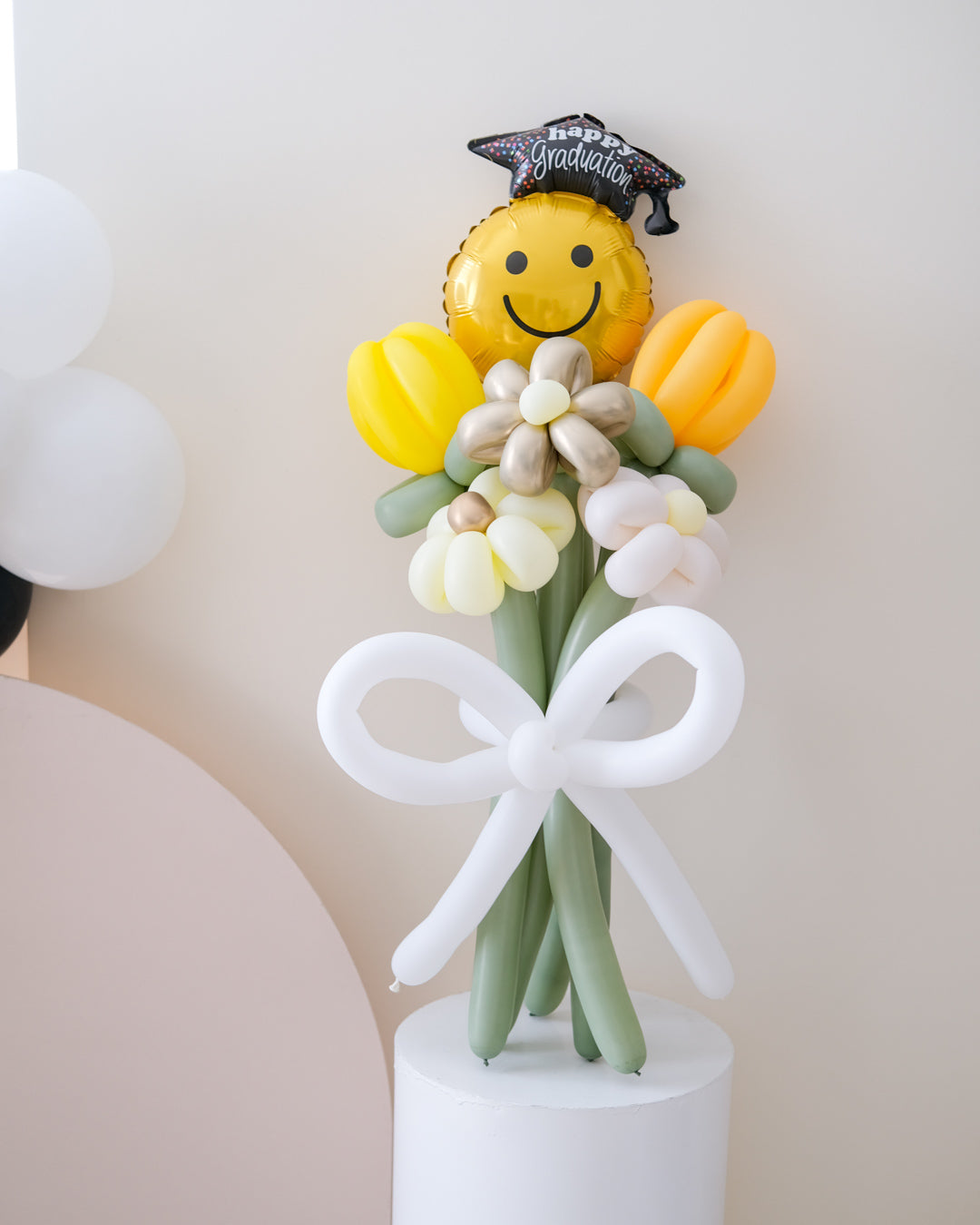 A balloon bouquet featuring a smiling face balloon, yellow and white flower-shaped balloons, and a balloon bow, standing on top of a column