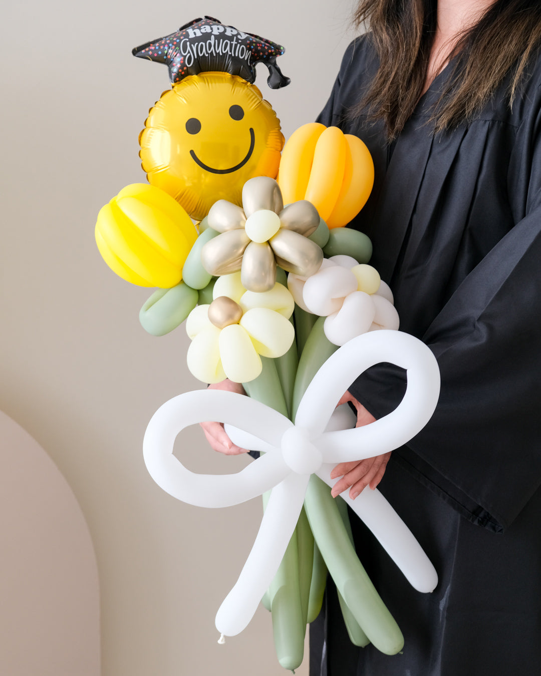 A balloon bouquet featuring a smiling face balloon, yellow and white flower-shaped balloons, and a balloon bow, held by a person in graduation attire.