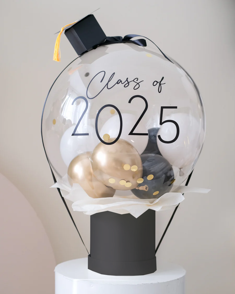 A decorative bubble balloon box with 'Class of 2025' printed on it, filled with multicolored balloons, mounted on a black stand.