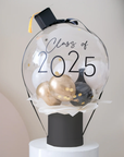 A decorative bubble balloon box with 'Class of 2025' printed on it, filled with multicolored balloons, mounted on a black stand.