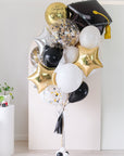 Graduation-themed balloons with a black cap, gold confetti, and foil stars with a tassel on a light background 