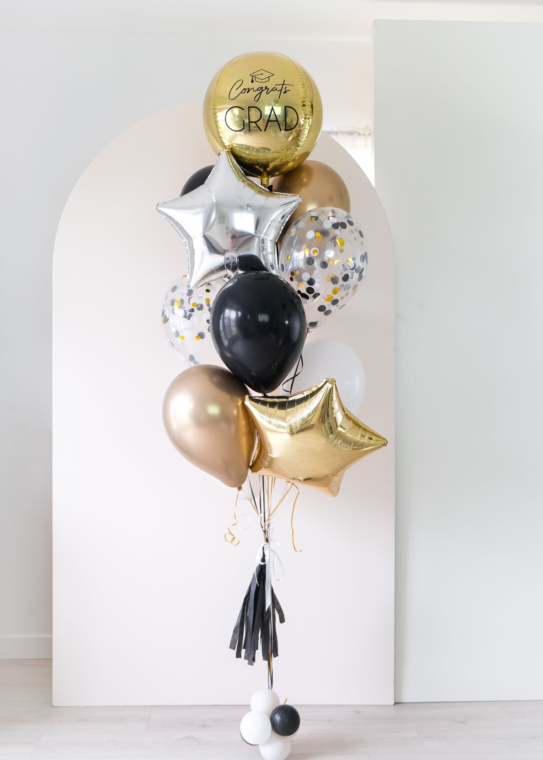 gold, black, white, cream, and silver graduation balloons in front of an arch backdrop
