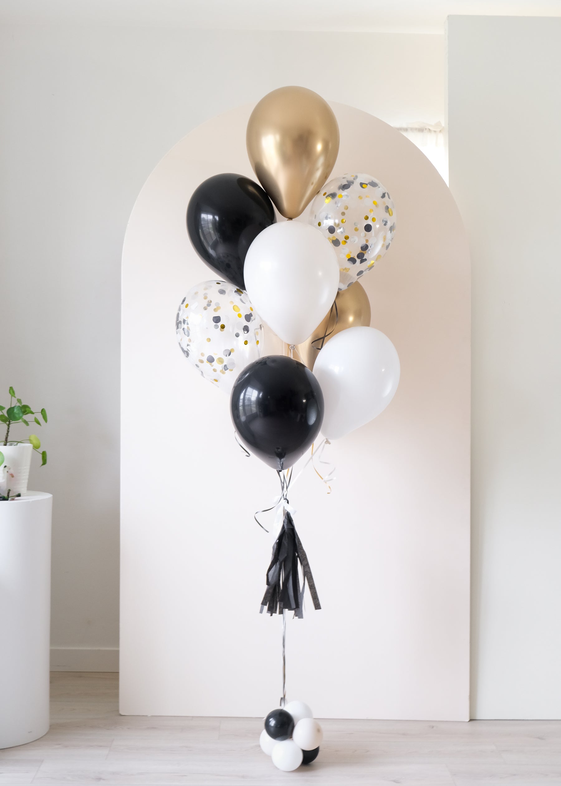 graduation balloon bouquet in black, white, cream. confetti-filled, and gold colors in front of an arch backdrop