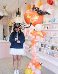 Person holding a spider balloon next to a halloween pumpkin balloon sculpture in a store setting