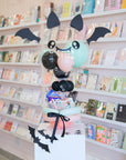 Decorative balloon arrangement with bat wings and a ghost head in front of a bookshelf.
