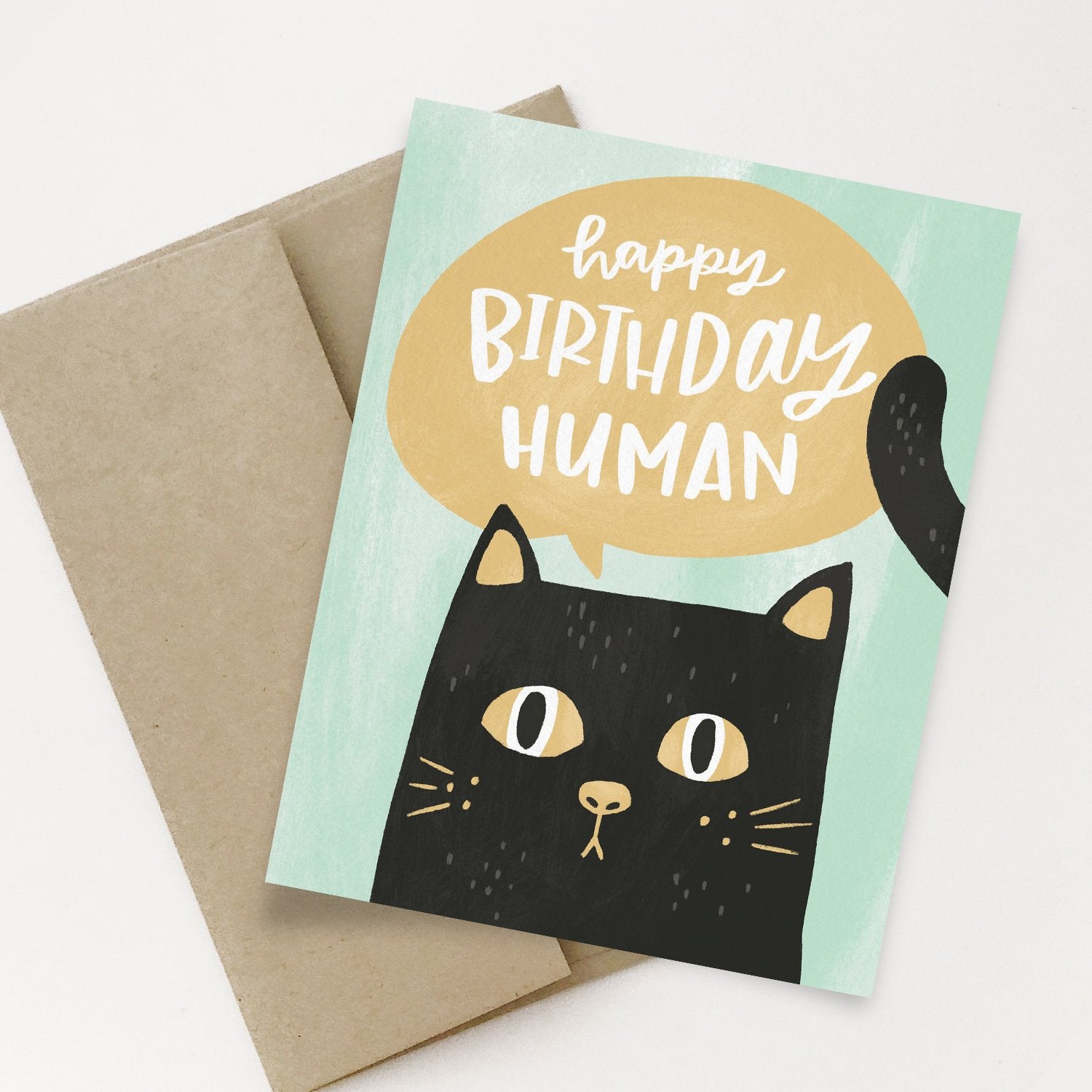 A card with a cat illustration and 'Happy Birthday Human' text, placed on top of a beige envelope.