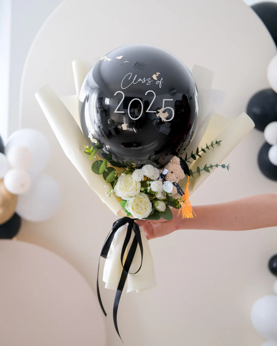 A hand holding a mini graduation bouquet with a large black balloon featuring 'Class 2025', wrapped with black and white paper, and decorated with white and yellow flowers and a small black bear figurine.