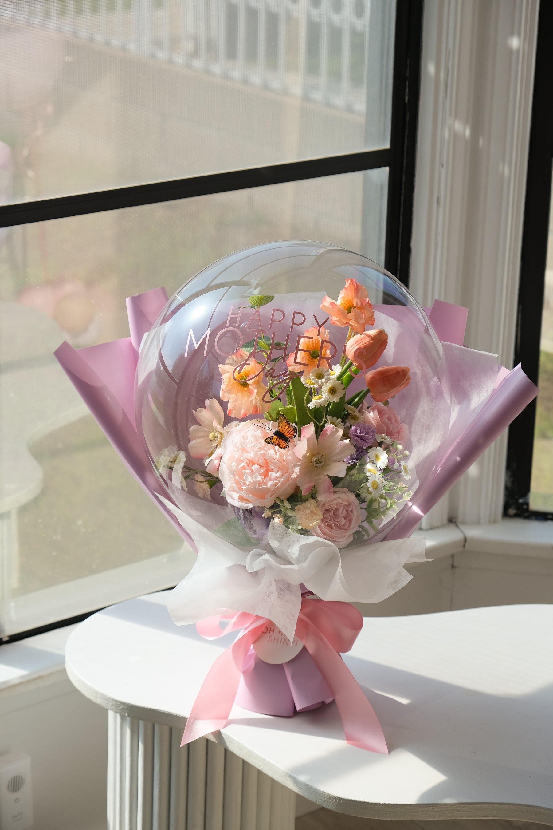 Bouquet of faux spring flowers in a transparent balloon with 'Happy Mother's Day' text, wrapped in lavender paper and ribbon by a window 