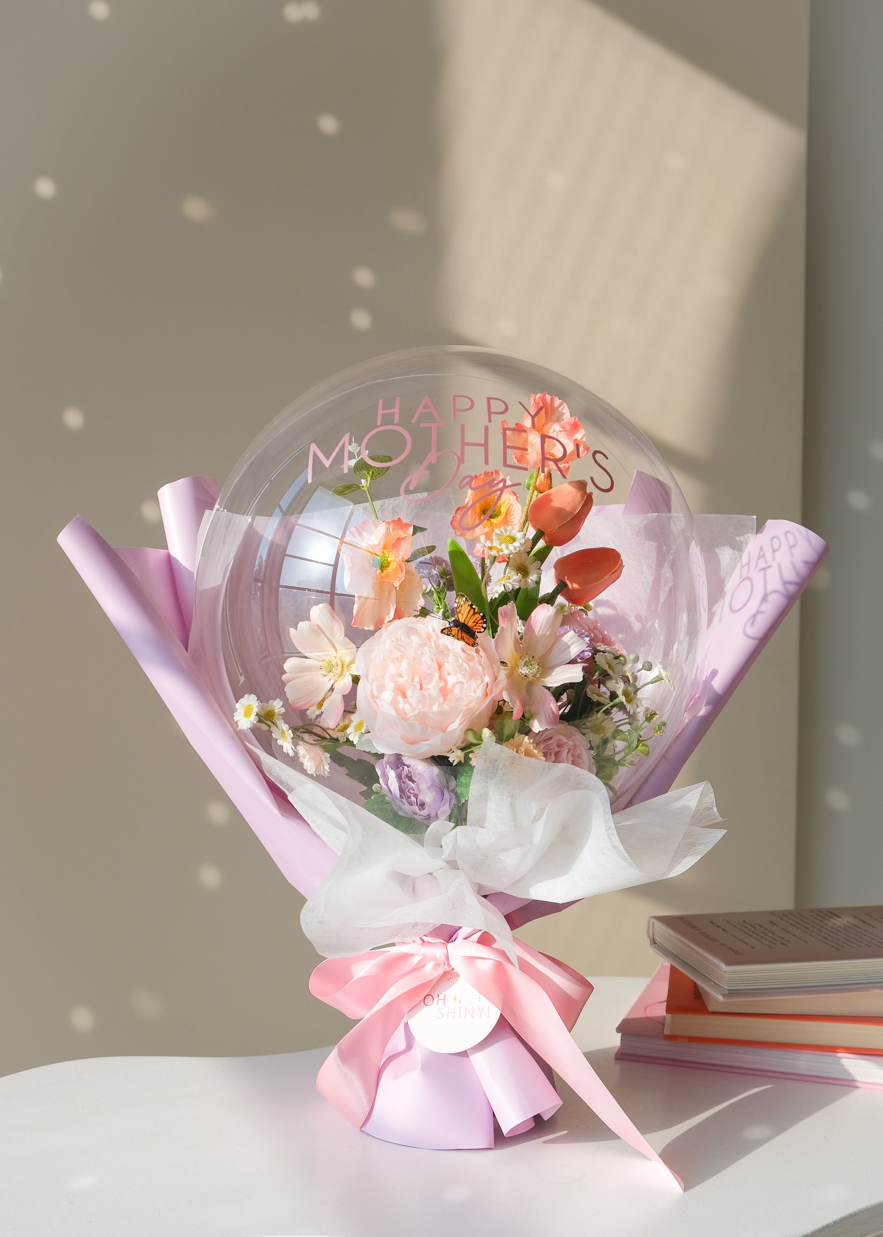 Bouquet of faux spring flowers in a transparent balloon with 'Happy Mother's Day' text, wrapped in lavender paper and ribbon 