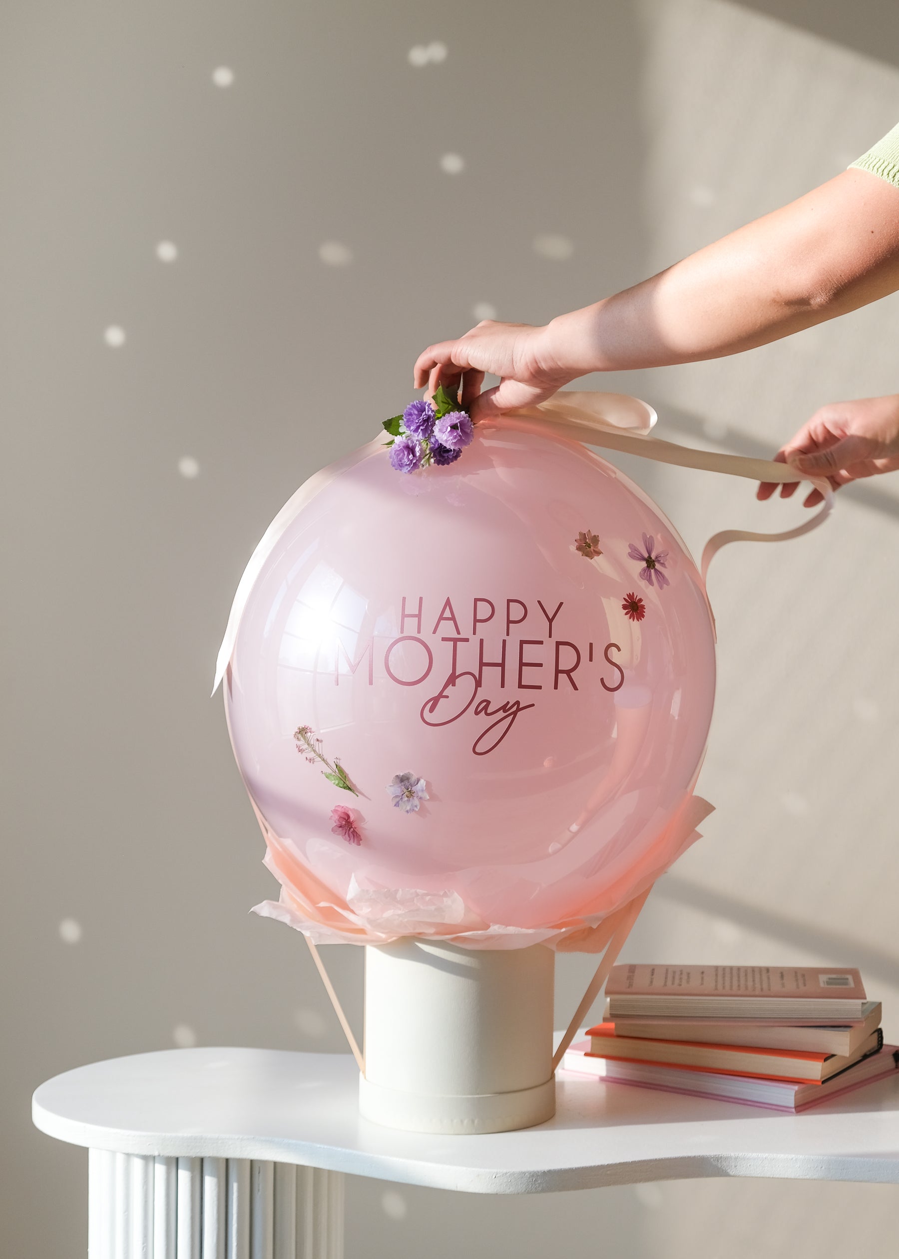 Hand unraveling ribbon from a pink "Happy Mother's Day" balloon on a box 