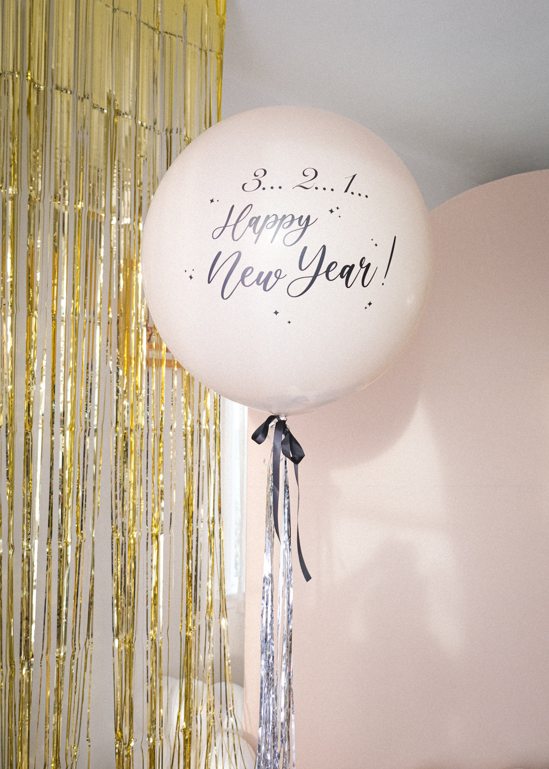 Cream round balloon with black bow and silver metallic tinsel. Text reads "3..2..1.. Happy new Year!" with gold tinsel and cream backdrop