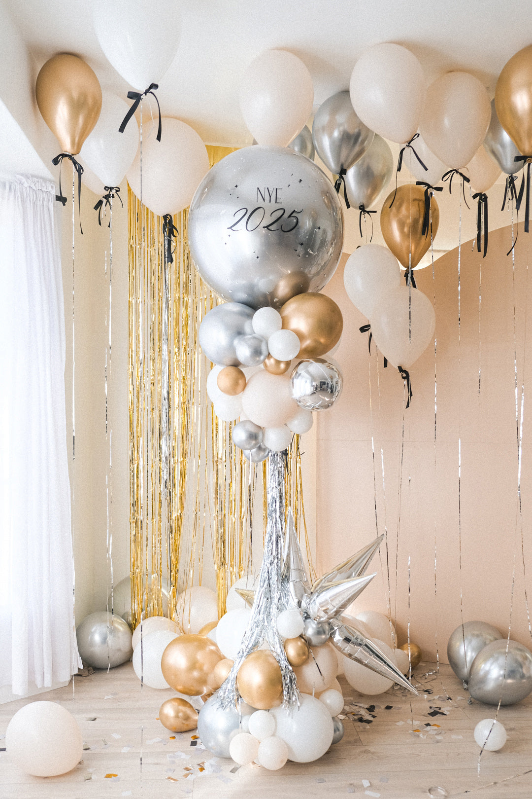 nye 2025 balloon tower with a giant round silver balloon and clusters of gold, silver and cream balloons underneath on a stand. Helium balloons with black bows floating on the ceiling.