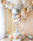 nye 2025 balloon tower with a giant round silver balloon and clusters of gold, silver and cream balloons underneath on a stand. Helium balloons with black bows floating on the ceiling.