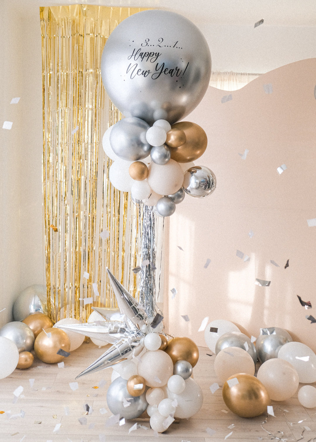 A round silver balloon with New Year text accompanied with smaller clusters of balloons underneath on a stand. Gold tinsel and cream backdrop in the background.
