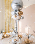 A round silver balloon with New Year text accompanied with smaller clusters of balloons underneath on a stand. Gold tinsel and cream backdrop in the background.