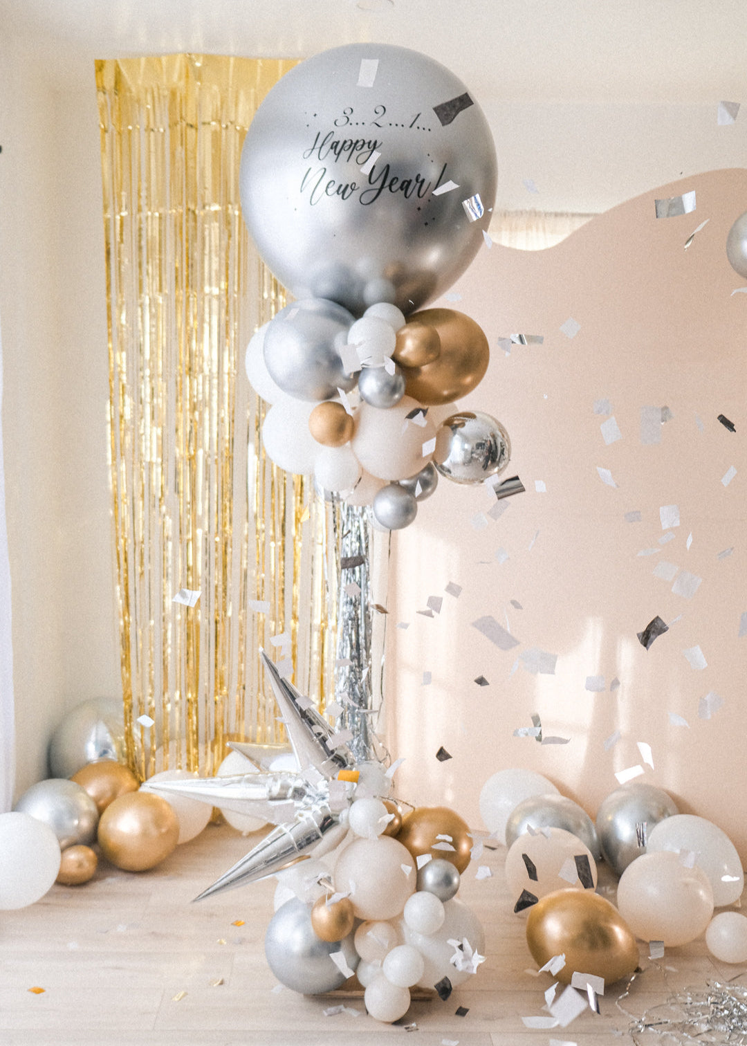 A round silver balloon with New Year text accompanied with smaller clusters of balloons underneath on a stand. Gold tinsel and cream backdrop in the background. Confetti accents are flying on the side.