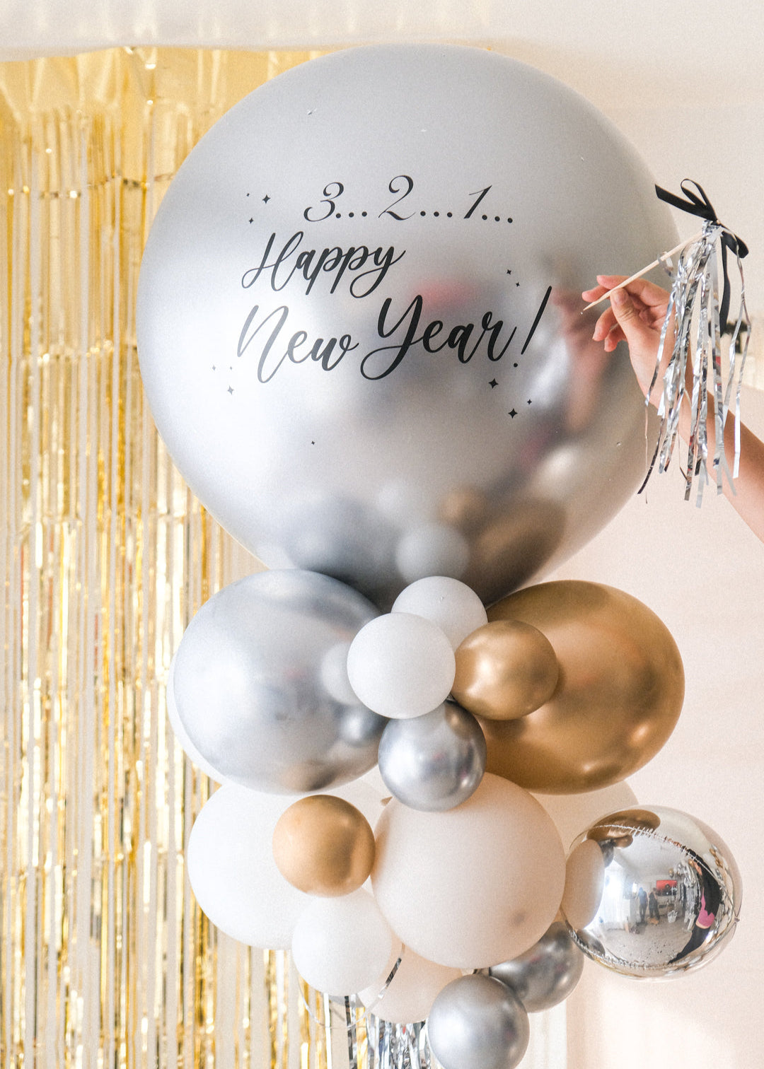 A close up shot of the round silver balloon with New Year text accompanied with smaller clusters of balloons underneath on a stand. Gold tinsel and cream backdrop in the background.
