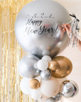 A close up shot of the round silver balloon with New Year text accompanied with smaller clusters of balloons underneath on a stand. Gold tinsel and cream backdrop in the background.