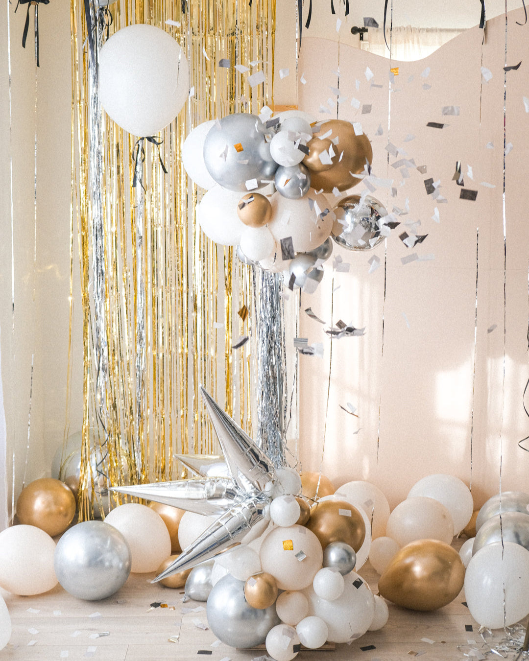 Confetti flying around from the popped round balloon accompanied with smaller clusters of balloons underneath on a stand. Gold tinsel and cream backdrop in the background.