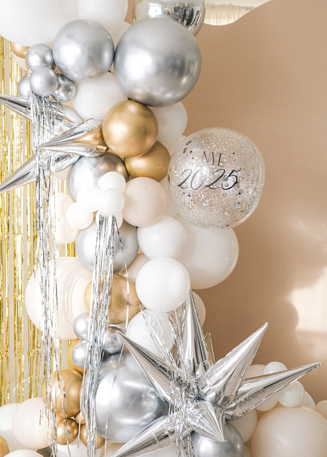 A balloon galrand in shiny silver, gold, white and creams with metallic tinsel and spikey balloon accents against a cream backdrop and gold tinsel curtain. A clear balloon that reads "NYE 2025"