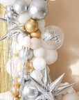 A balloon galrand in shiny silver, gold, white and creams with metallic tinsel and spikey balloon accents against a cream backdrop and gold tinsel curtain. A clear balloon that reads "NYE 2025"