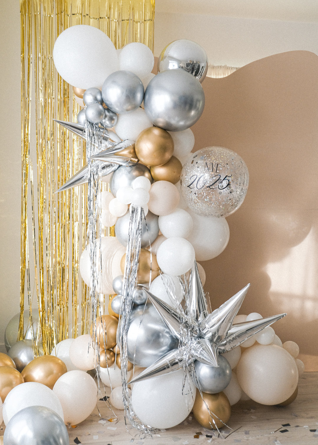 A balloon galrand in shiny silver, gold, white and creams with metallic tinsel and spikey balloon accents against a cream backdrop and gold tinsel curtain.