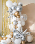 A balloon galrand in shiny silver, gold, white and creams with metallic tinsel and spikey balloon accents against a cream backdrop and gold tinsel curtain.