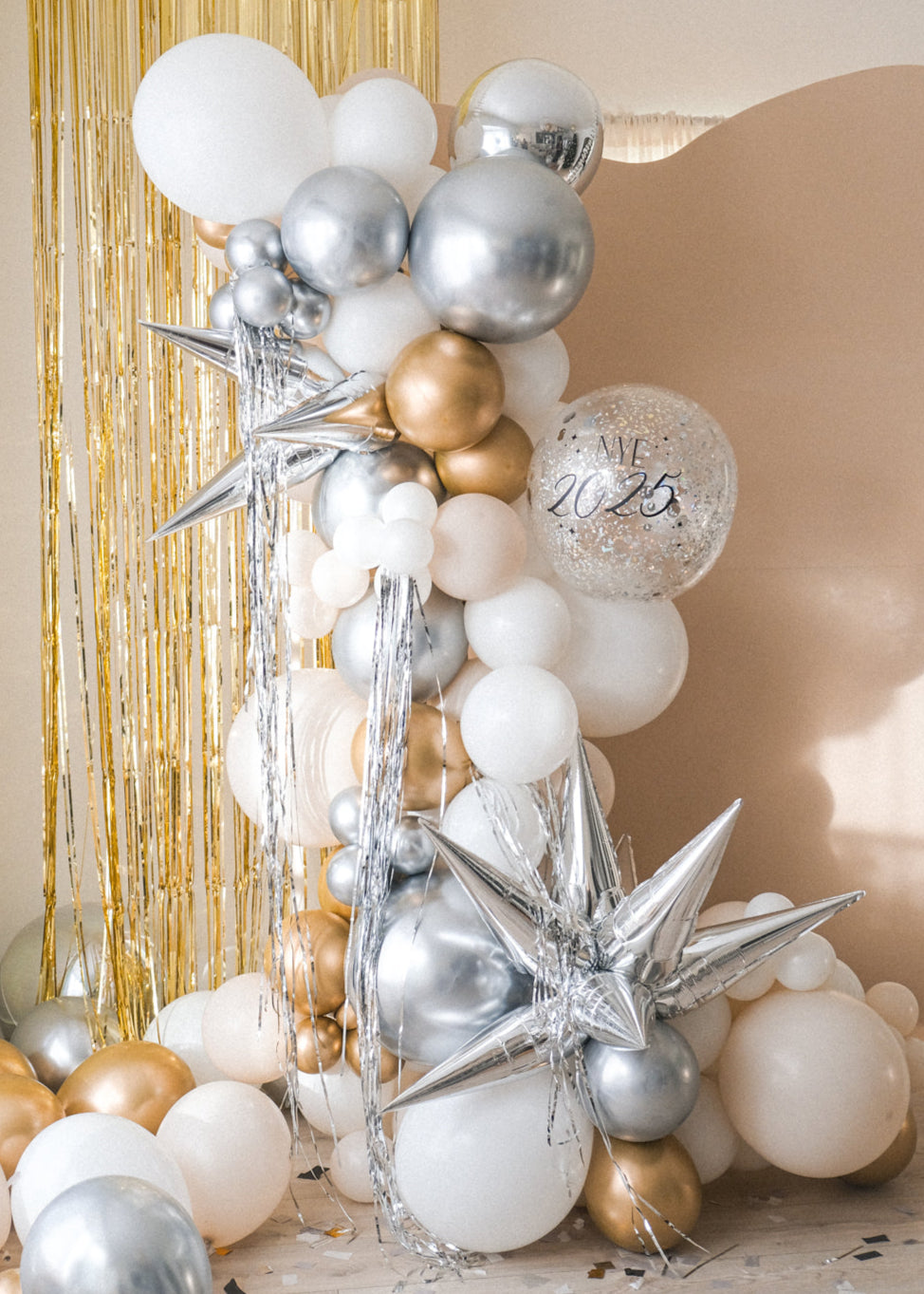 A balloon galrand in shiny silver, gold, white and creams with metallic tinsel and spikey balloon accents against a cream backdrop and gold tinsel curtain