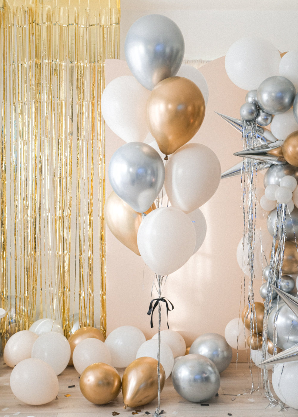 A larger bouquet of balloons in gold, silver, cream, and white colors with sparkly silver tinsel strings, displayed indoors with a glittery backdrop.