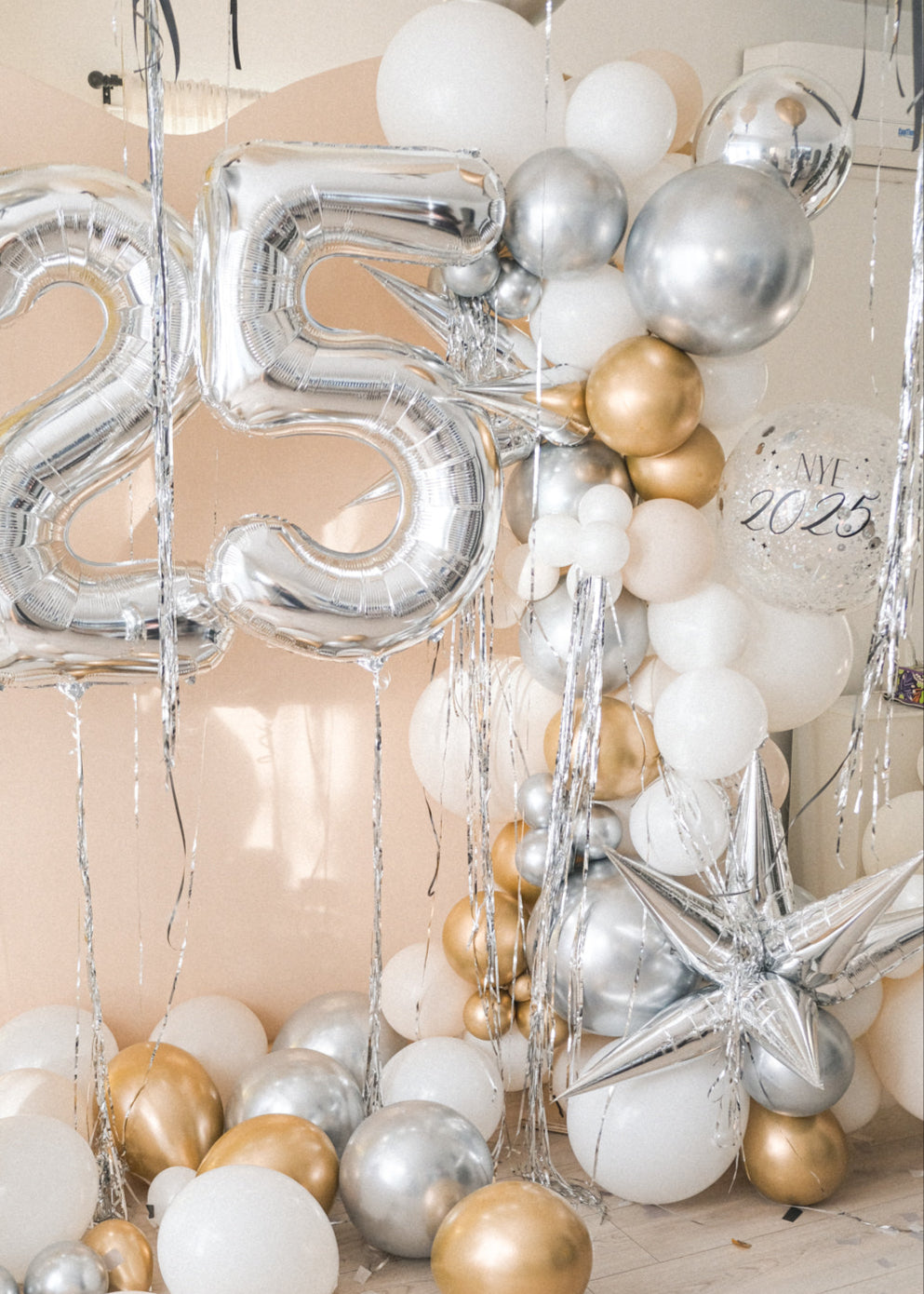 A balloon galrand in shiny silver, gold, white and creams with metallic tinsel and spikey balloon accents against a cream backdrop and silver numbers 25 to the left