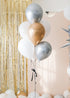 A bouquet of round balloons in gold, silver, cream, and white colors with sparkly silver tinsel strings, displayed indoors with a glittery backdrop.