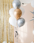 A bouquet of round balloons in gold, silver, cream, and white colors with sparkly silver tinsel strings, displayed indoors with a glittery backdrop.