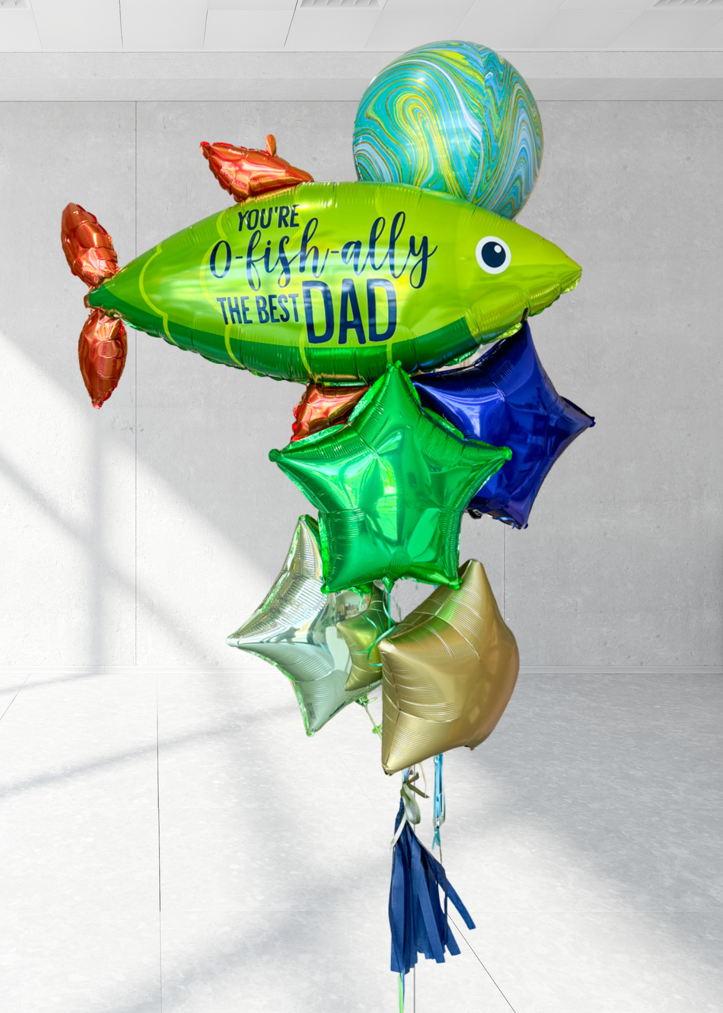 A bunch of colorful balloons including a large green one with 'You-O-Fish-ally The Best Dad' printed on it, along with various other balloons in shades of blue and red.