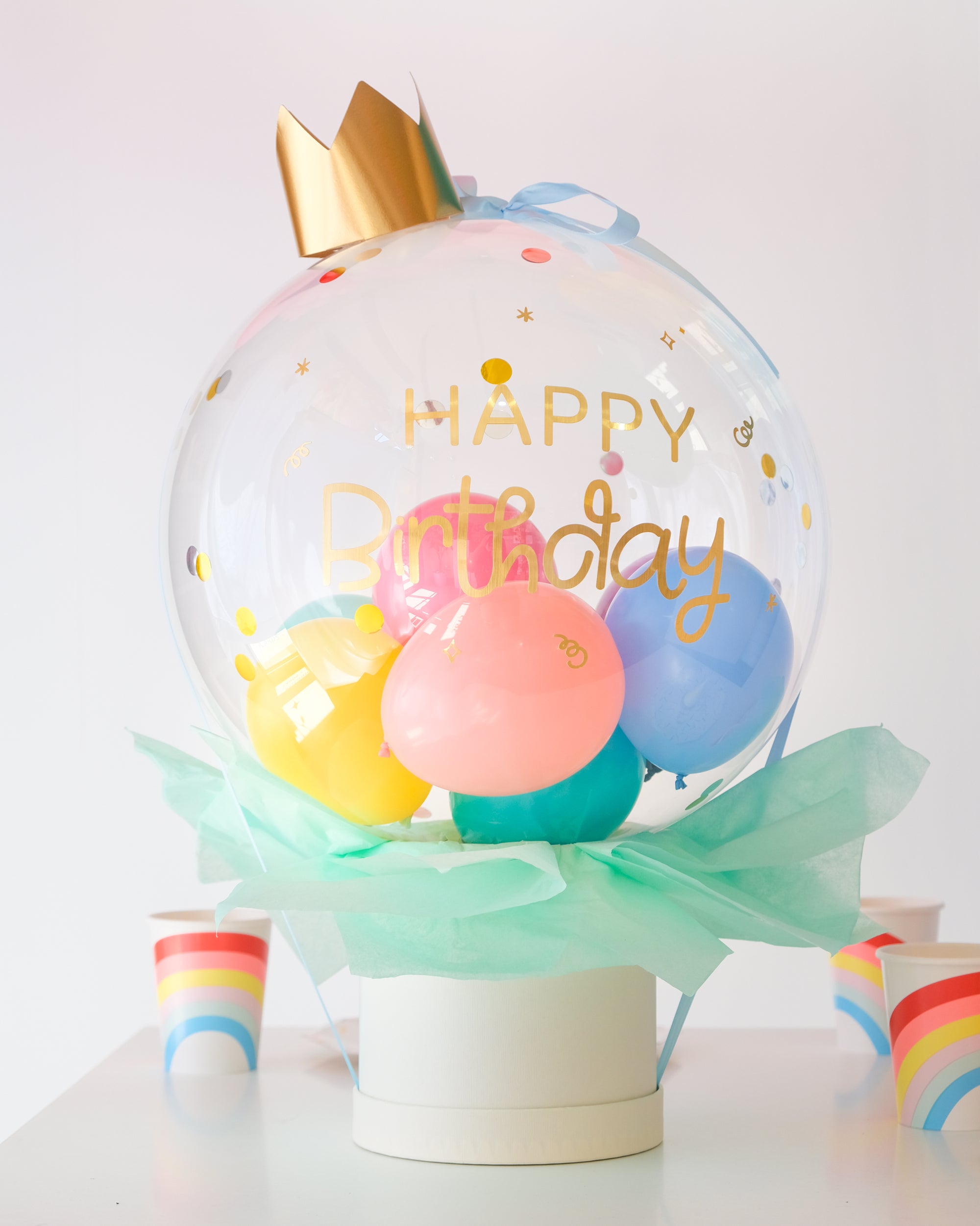A clear bubble box with a crown on top, decorated with colorful balloons and confetti, and labeled 'Happy Birthday'. Cups near the box on the table.